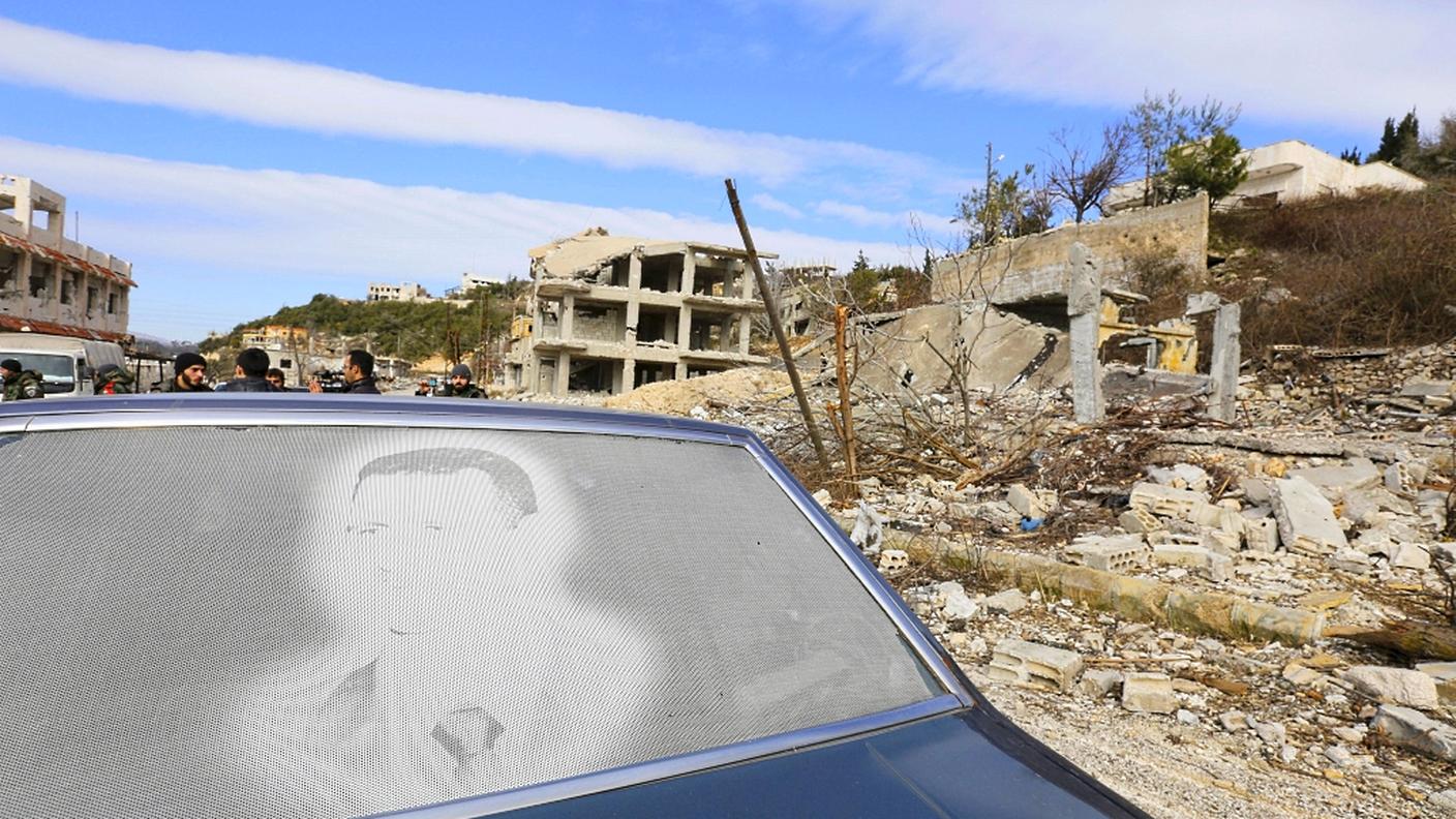 Un ritratto di Bashar al Assad; sullo sfondo, un paesaggio siriano colpito dalle bombe
